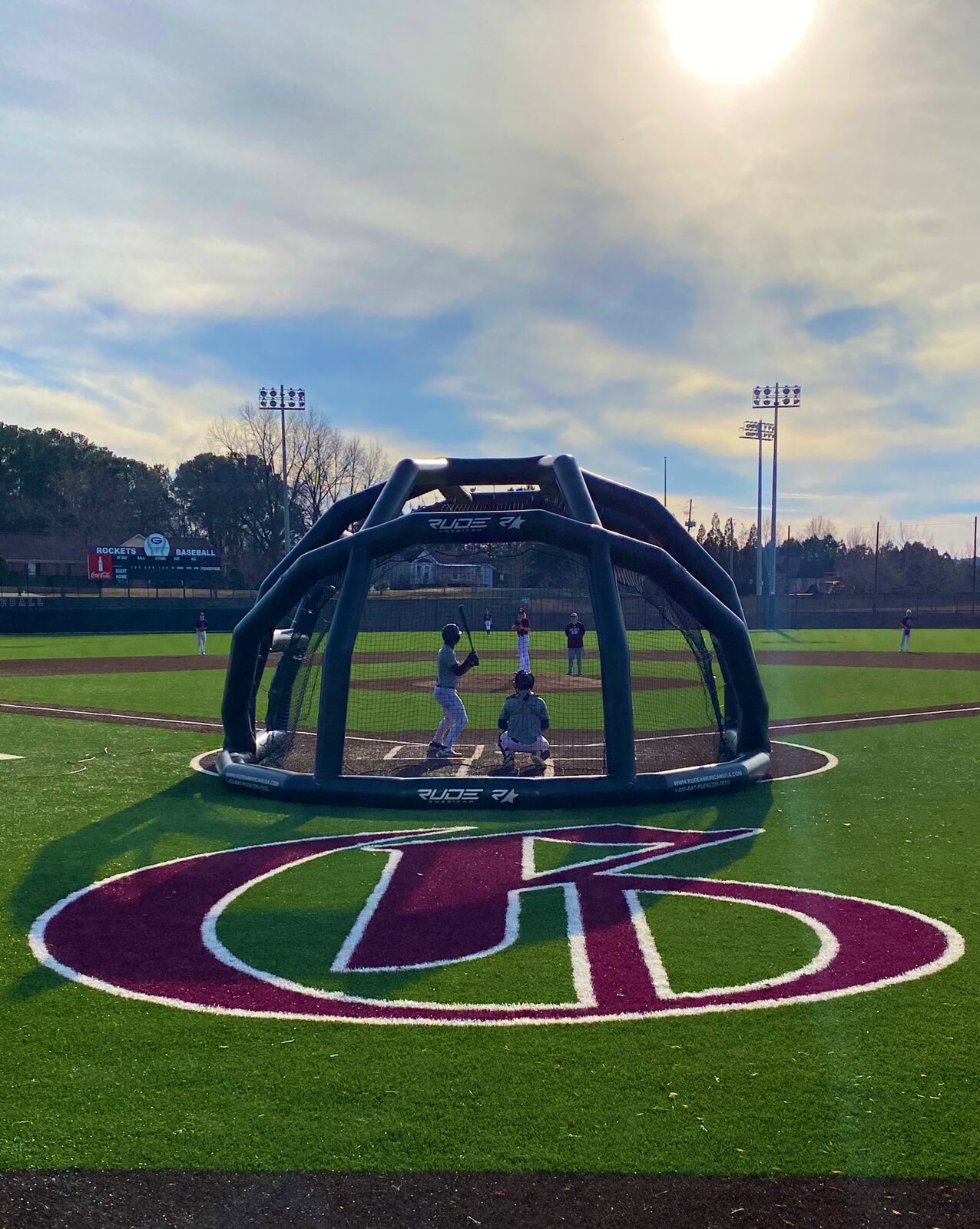 Batting practice in the portable cage at home plate, with the maroon R logo painted on the turf and the sun over the outfield.