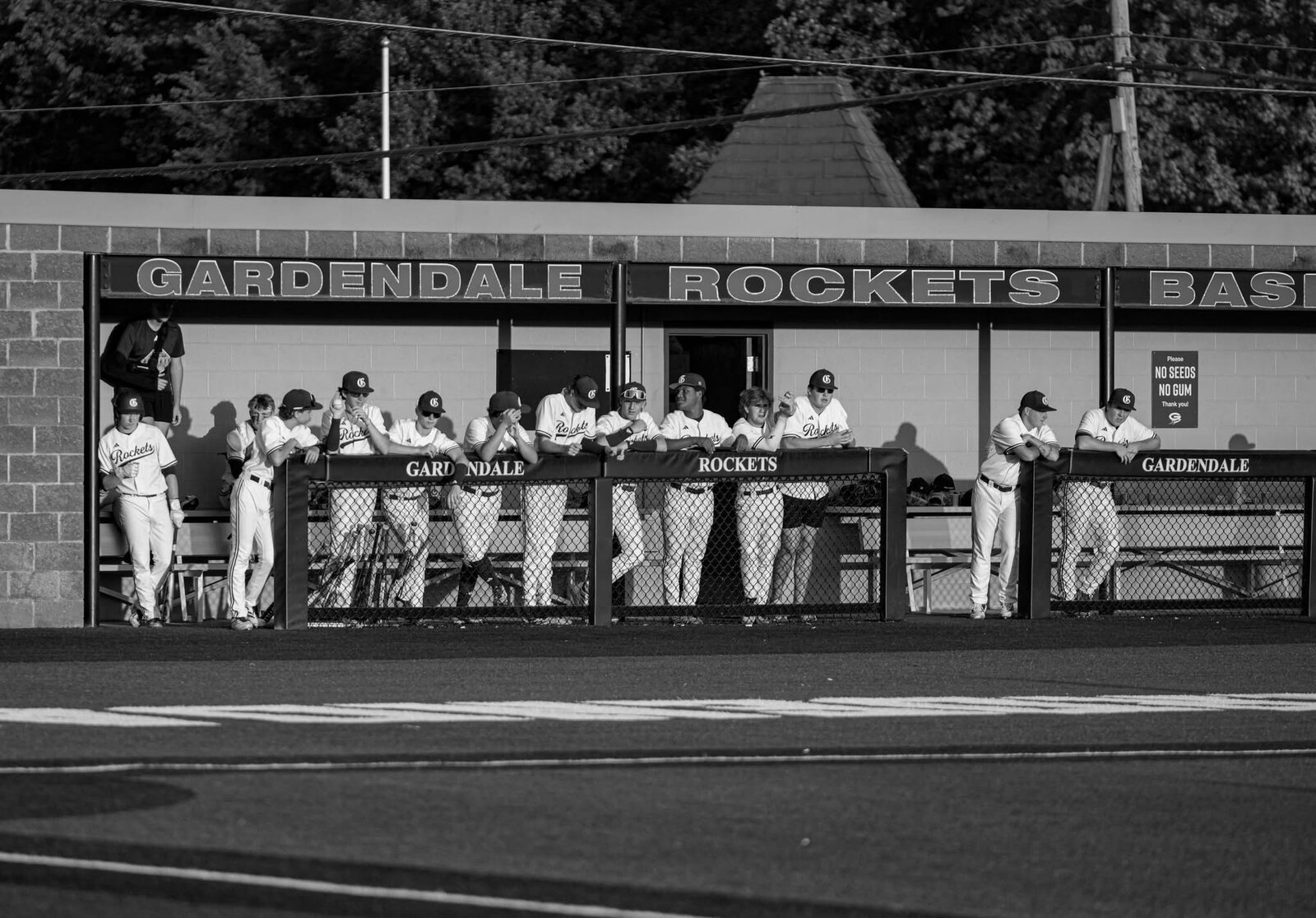 Players leaning on the dugout rail under the Gardendale Rockets Baseball sign, in black and white.